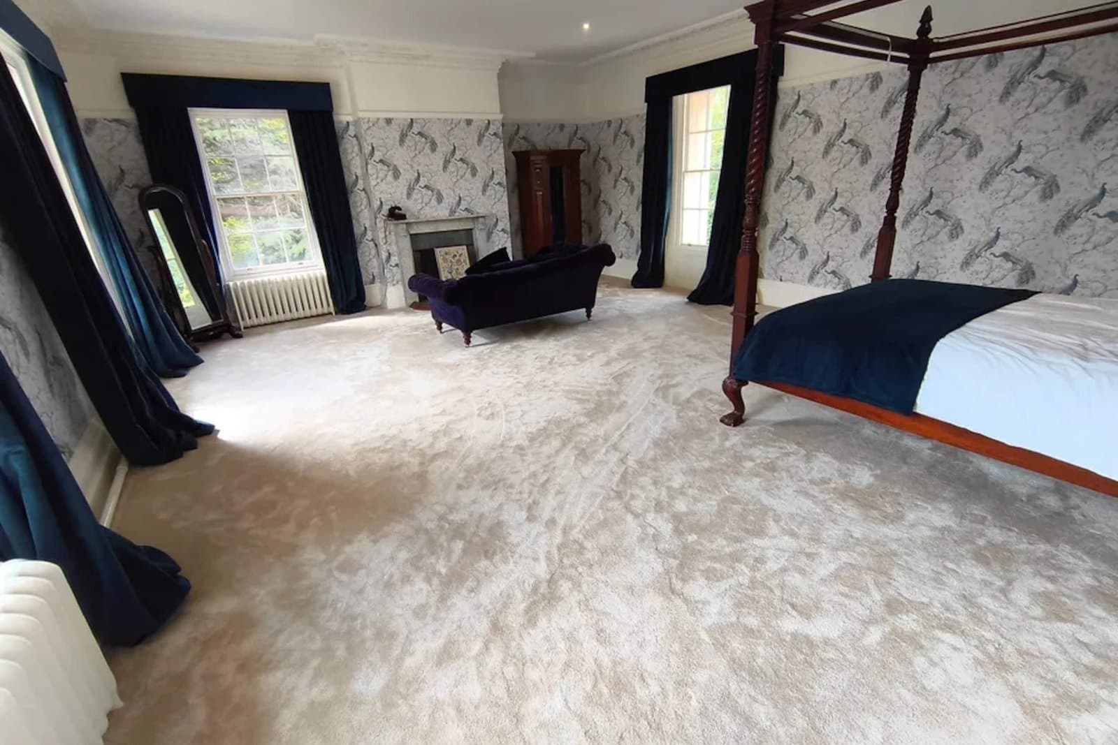 A four-poster bedroom with patterned wallpaper, navy velvet curtains, a chaise longue and a fireplace