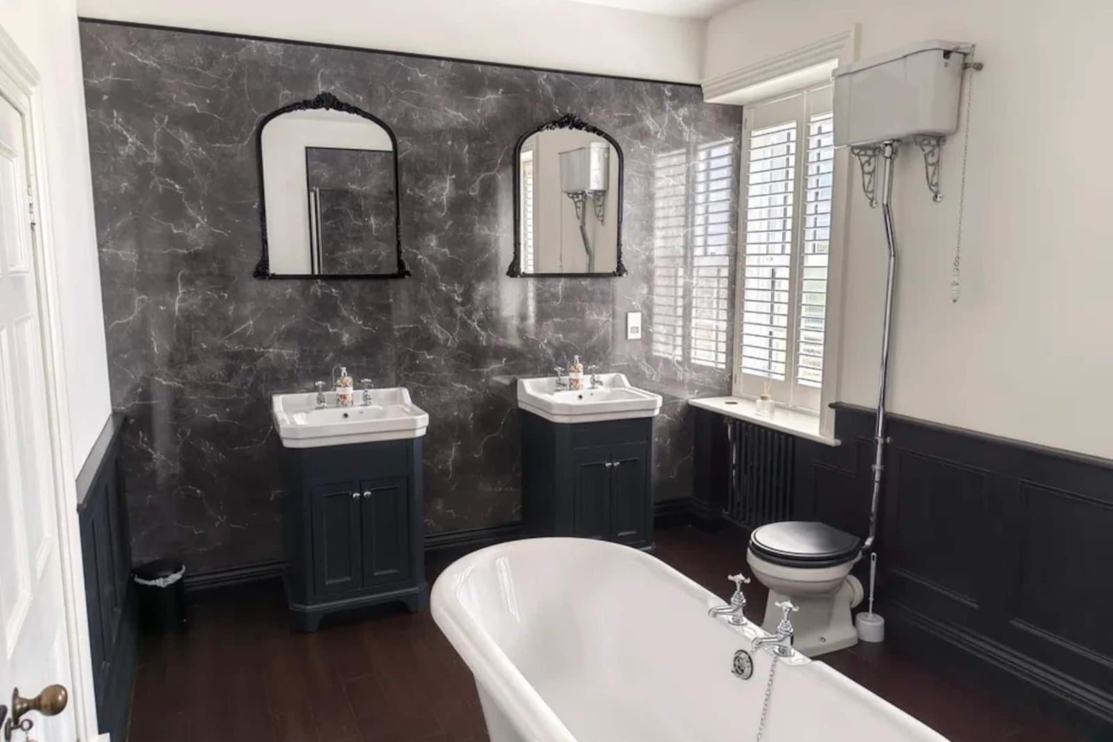A bathroom with a freestanding roll-top bath, twin basins, arched mirrors and dark panelled walls